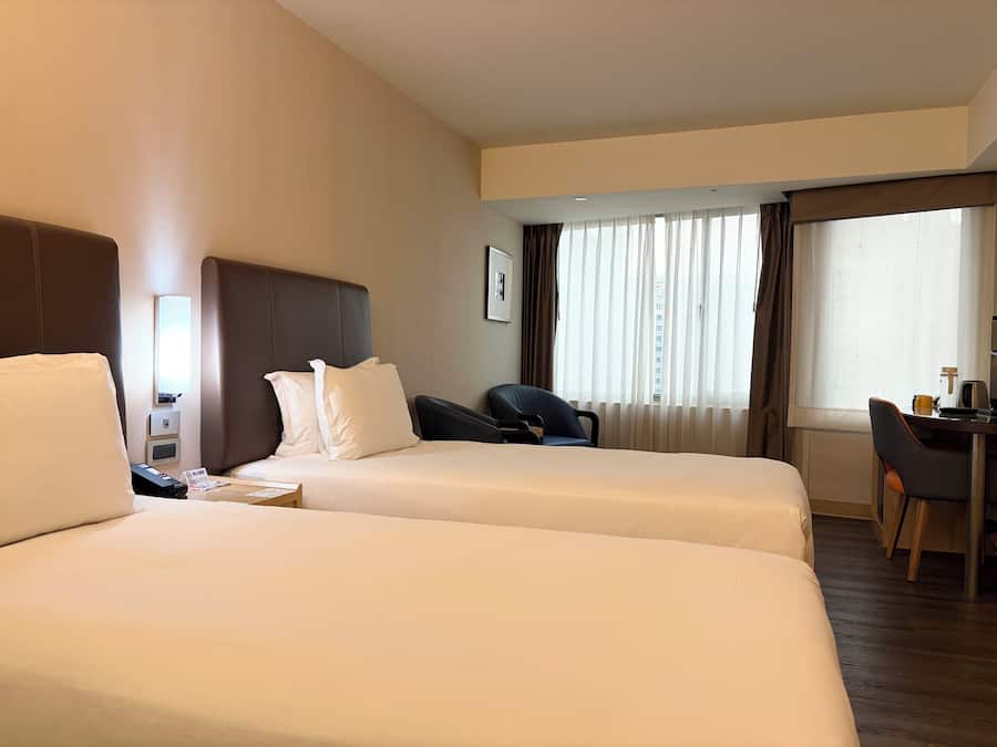Ein Hotelzimmer mit zwei Betten, einem Schreibtisch, einem Stuhl und einem fensterbepflanzten Fenster.