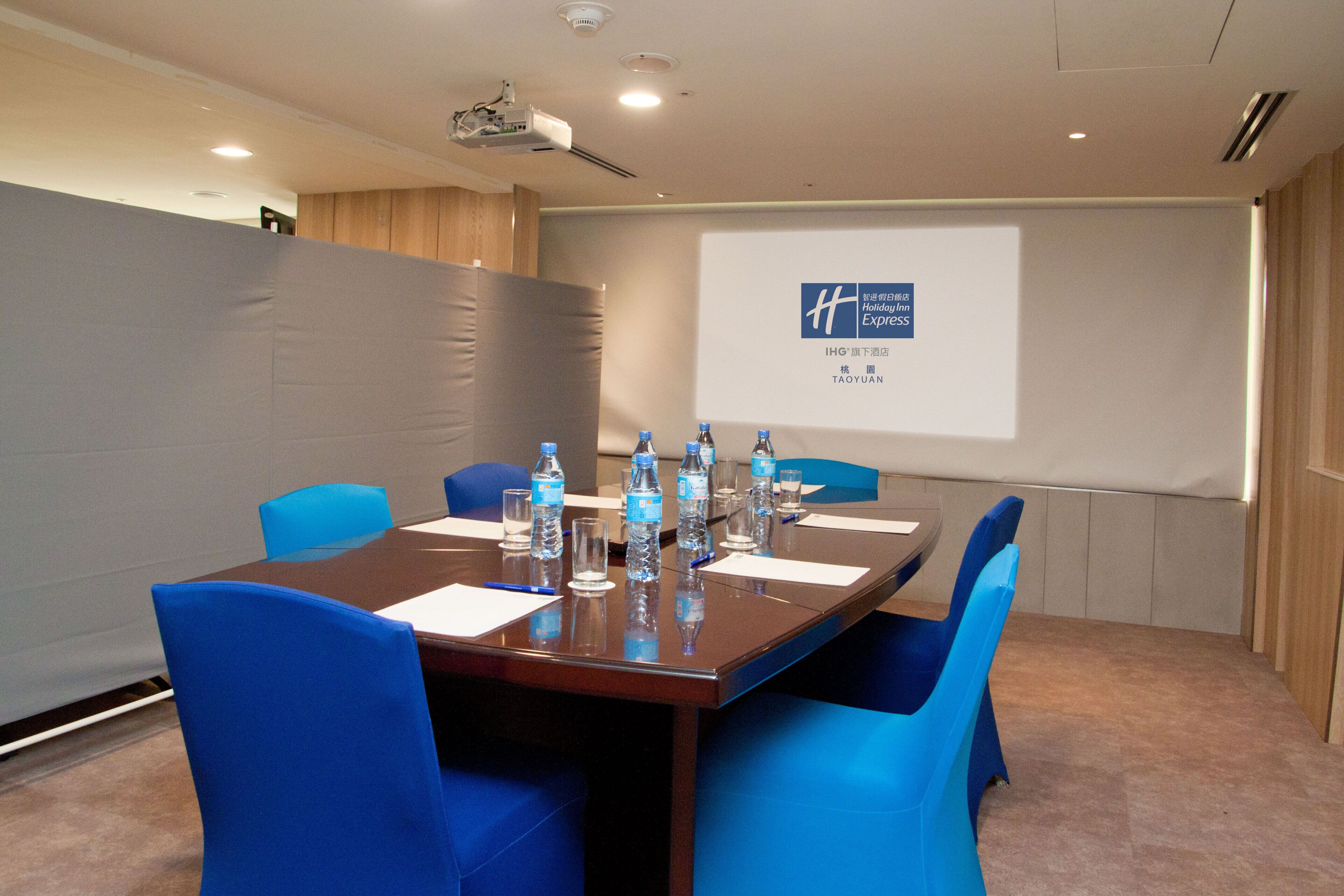 A conference room with a long table, blue chairs, water bottles, and a projector screen displaying a hotel name.