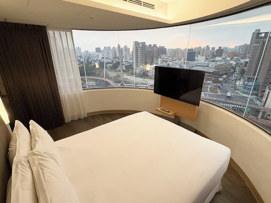 Ein Hotelzimmer mit einem großen Bett, einem flachbildfernseher an der Wand und Blick auf eine Stadtlandschaft durch ein großes Fenster.