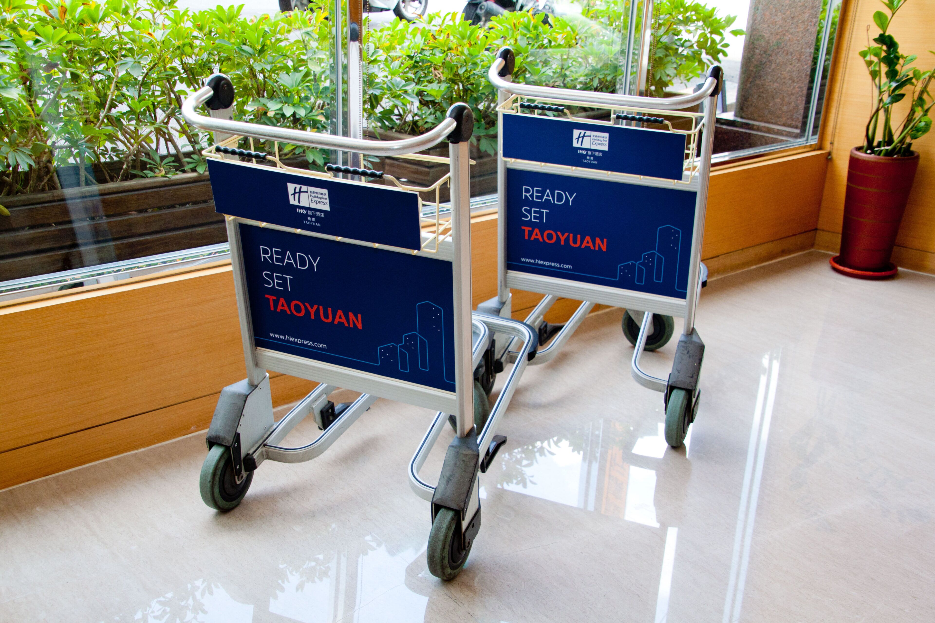 Zwei Einkaufswagen mit der Aufschrift 'READY SET TAOYUAN', platziert in einem Innenbereich mit spiegelndem Boden und Blumentöpfen im Hintergrund.