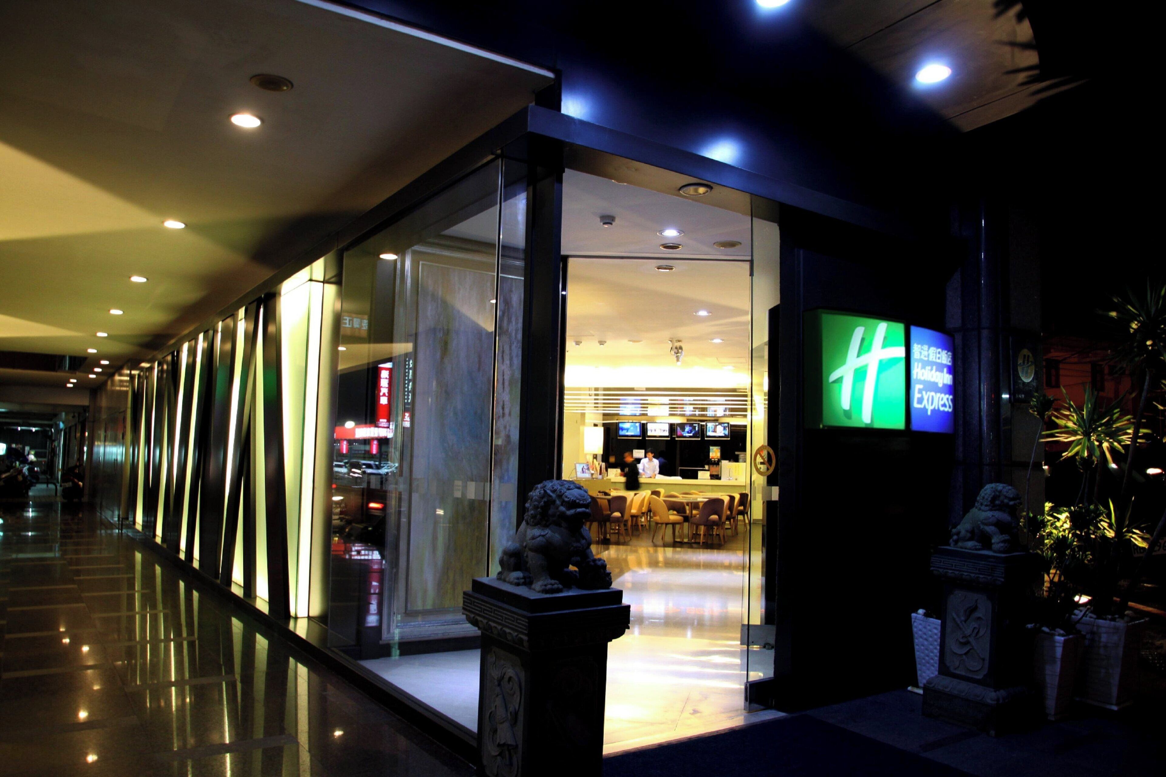 A modern hotel entrance with glass doors, a reception desk, and a digital signboard.