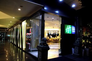 A modern hotel entrance with glass doors, a reception desk, and a digital signboard.