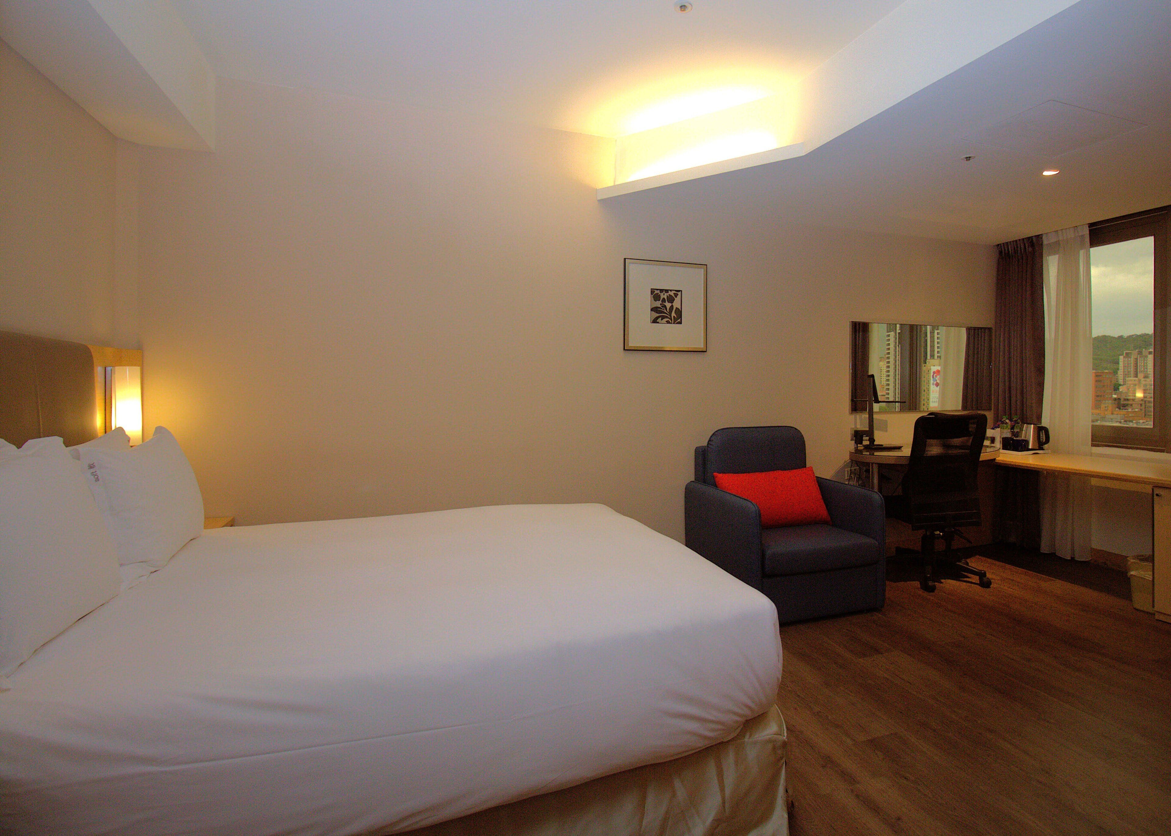 A hotel room with a large bed, a desk with a computer, and a chair with a red pillow.