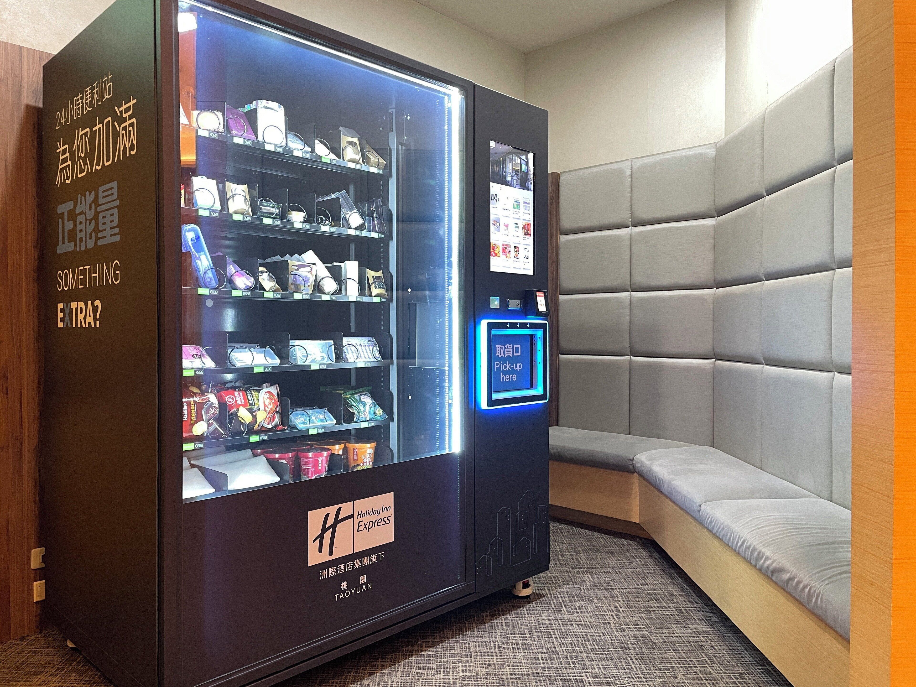 A Holiday Inn Express vending machine stocked with snacks and beverages, located in a corner with a cushioned seating area.