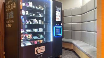 A Holiday Inn Express vending machine stocked with snacks and beverages, located in a corner with a cushioned seating area.