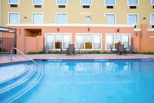 Piscina com borda azulejada, rodeada por um pátio de tijolos. Há cadeiras e mesas para hóspedes, e um edifício com várias janelas ao fundo.