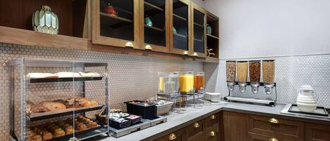Una barra de desayuno con pasteles, una estación de café y una vitrina con productos de panadería.