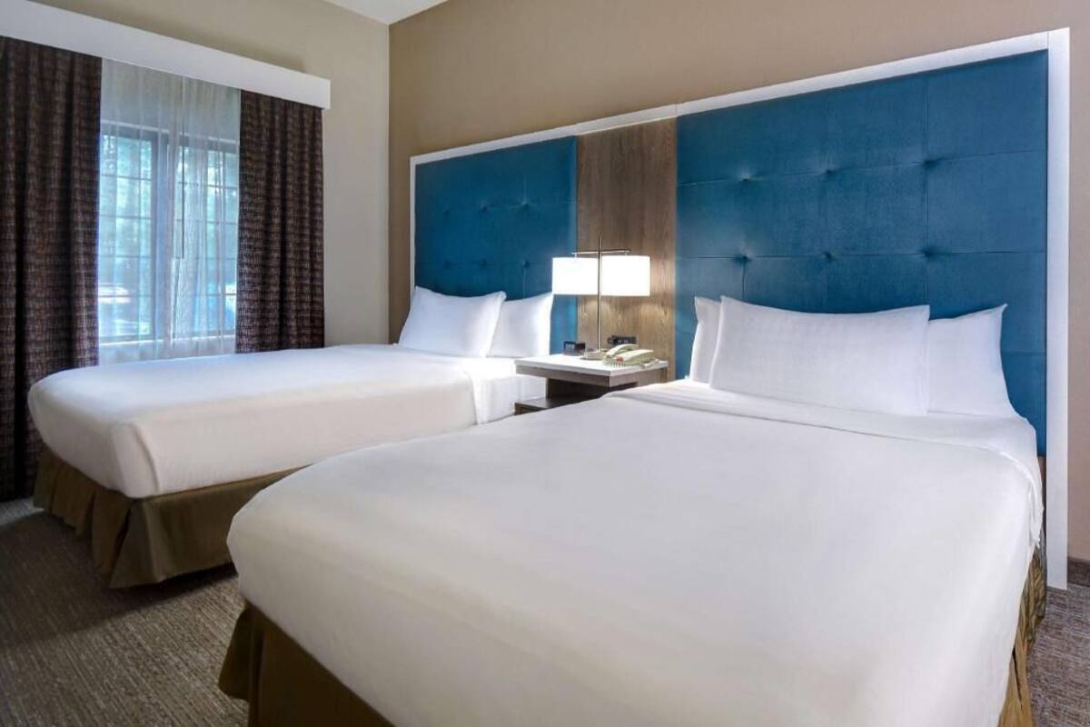 A hotel room with two beds, a large blue headboard, a bedside table with a lamp, and a window with curtains.