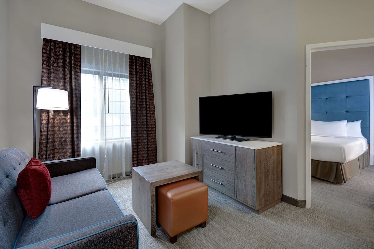 A hotel room with a sofa, a television, a small wooden cabinet, and a bed with a blue headboard.