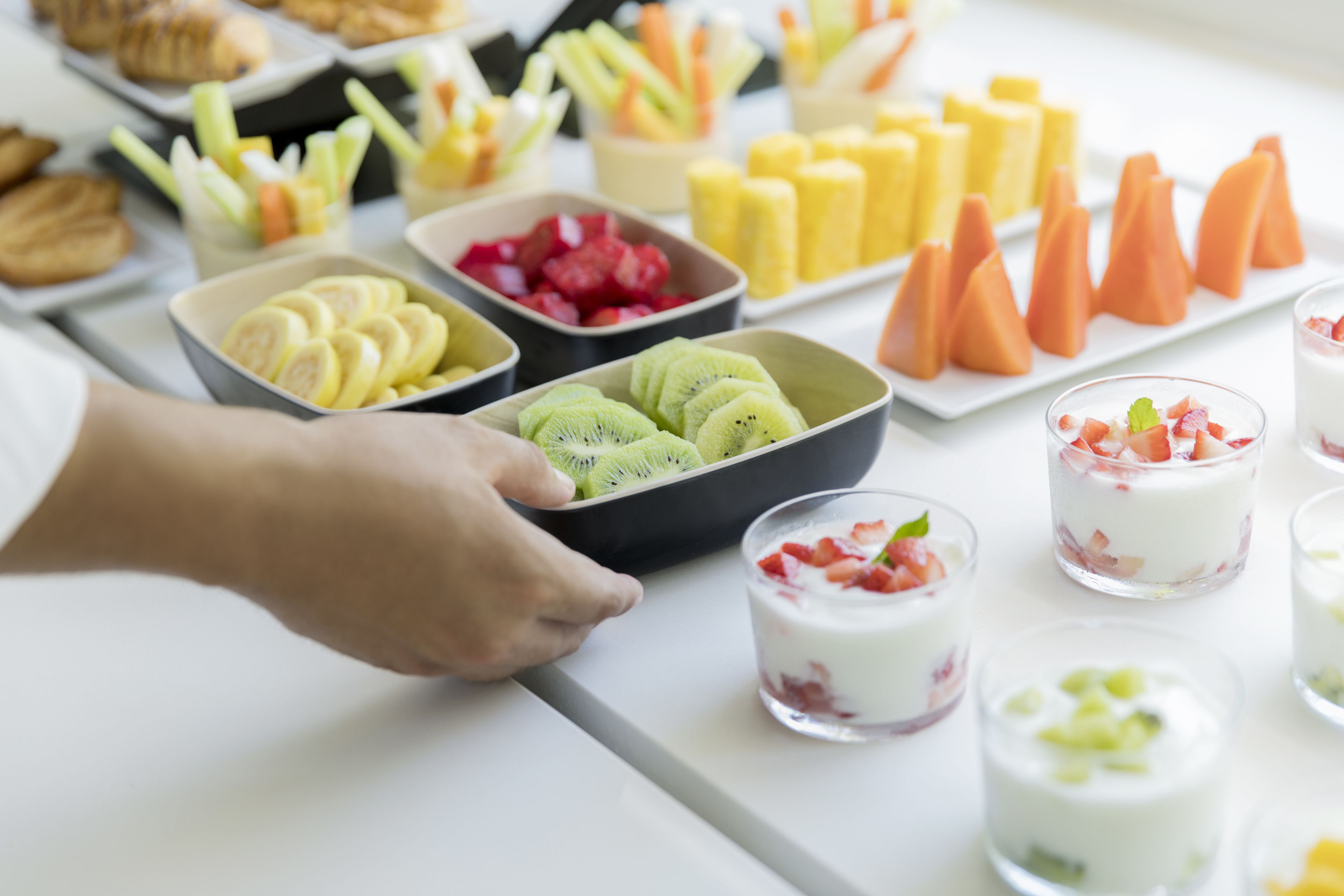 Forskellige spisemuligheder, herunder skåret frugt, yoghurt-parfaitter og kager, anrettet på et bord.