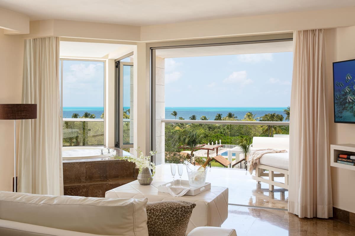 Quarto de hotel moderno com varanda e vista para o oceano, área de estar com mesa e uma grande janela com cortinas.