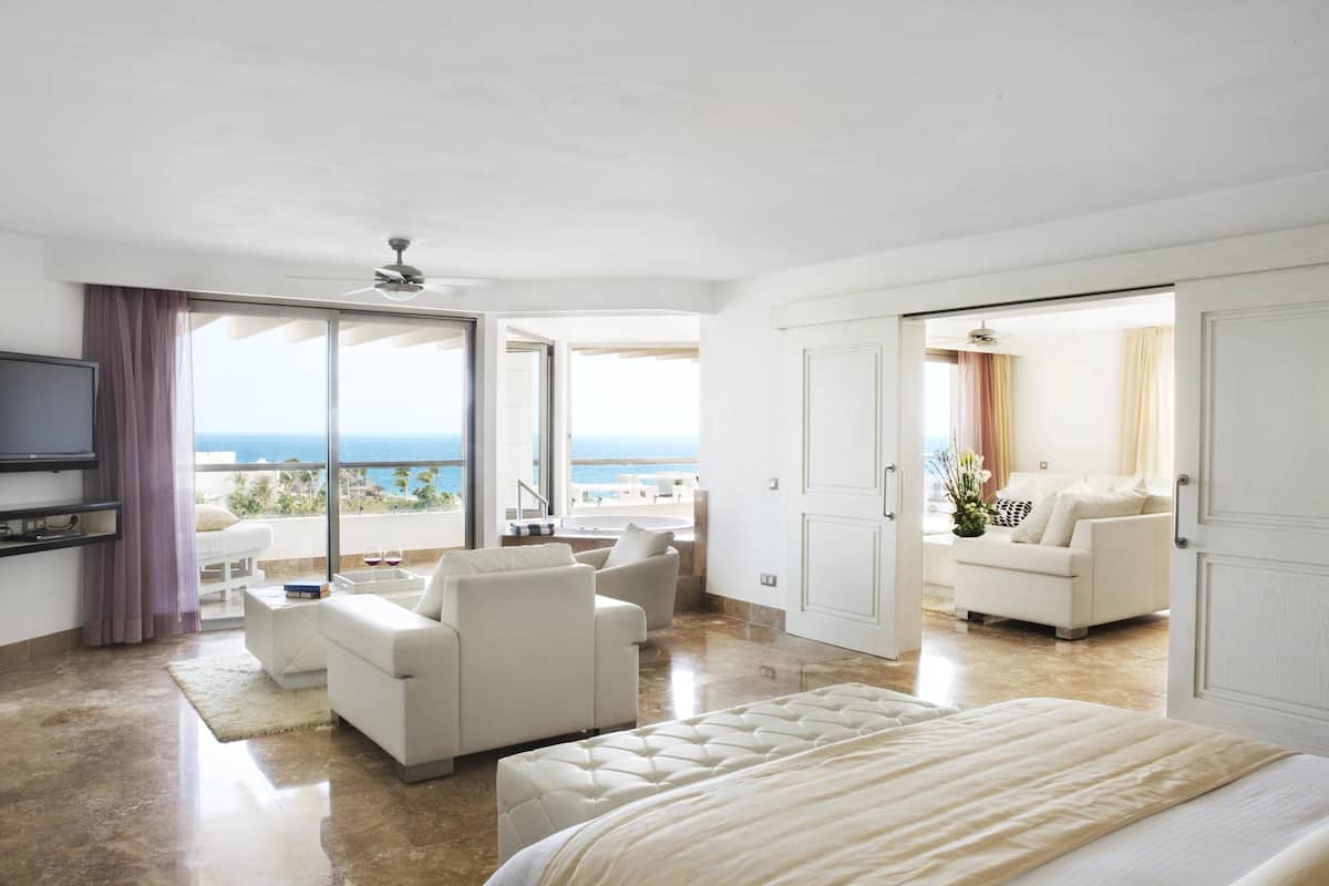 Quarto espaçoso com uma cama grande, área de estar com sofá e poltronas, e varanda com vista para o mar.