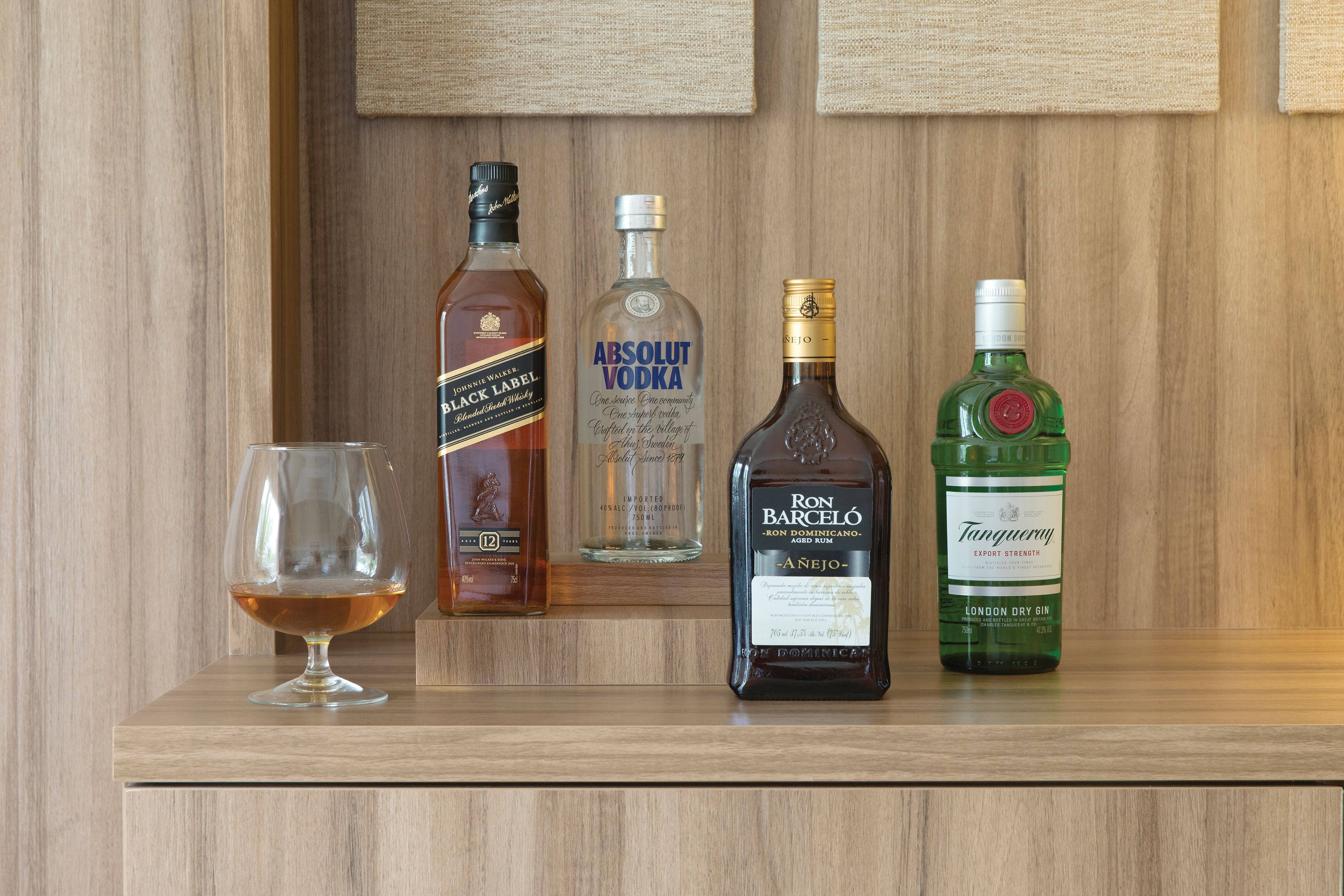 A wooden shelf with four bottles of alcohol and a glass.