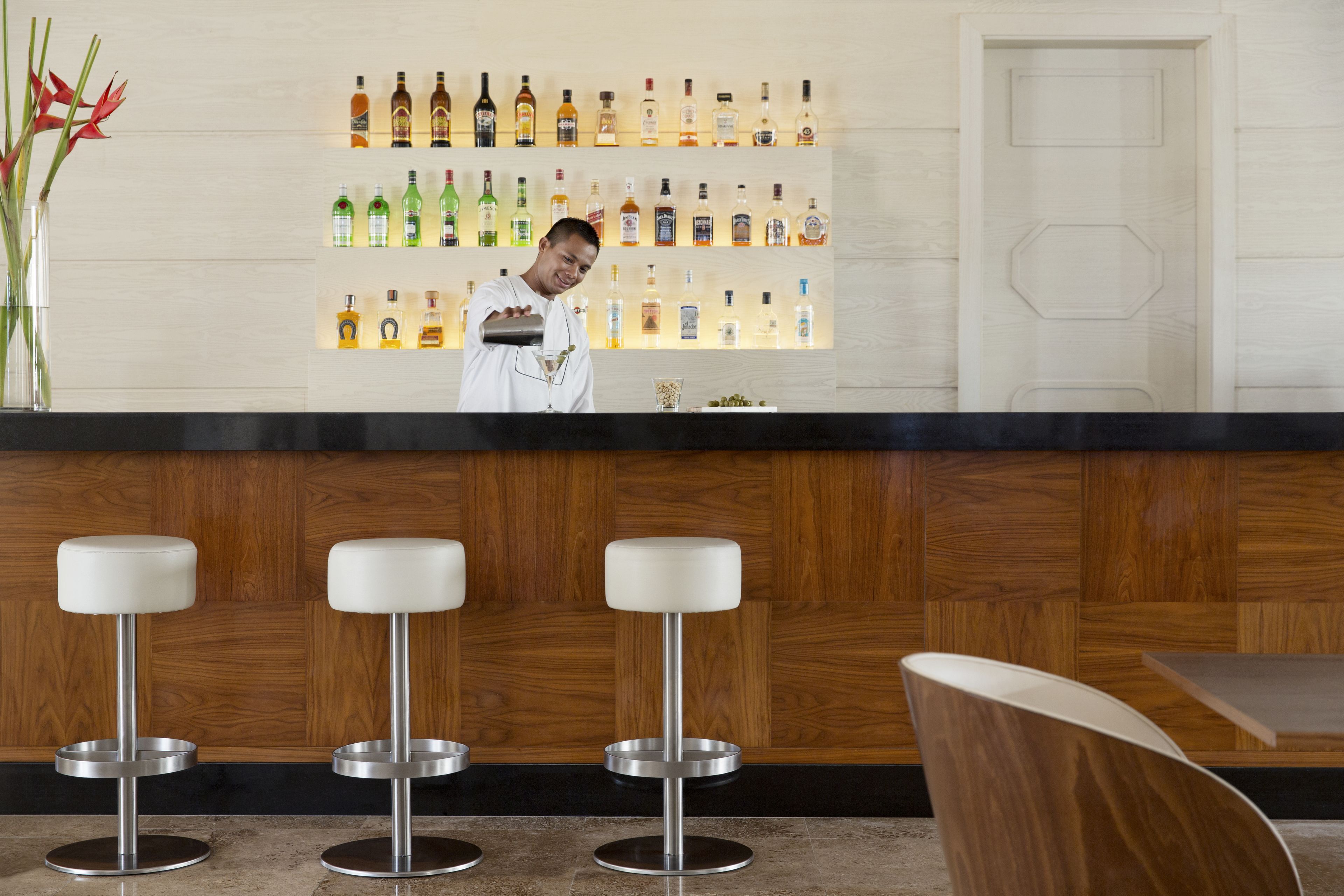 Uma área de bar com um bartender, uma parede de madeira com garrafas de bebidas e três banquetas brancas.