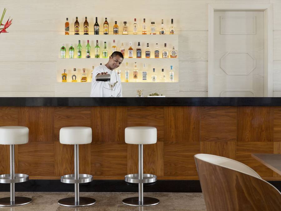 Uma área de bar com um bartender, uma parede de madeira com garrafas de bebidas e três banquetas brancas.
