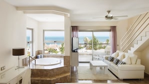 Ein modernes Hotelzimmer mit einem großen Bett, einer Sitzecke mit einem weißen Sofa, einem Esstisch, einer Badewanne und einem Balkon mit Meerblick.