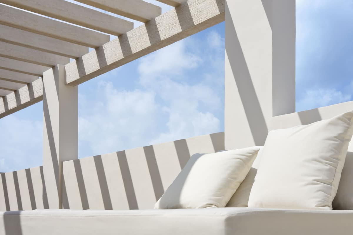 Uma área de estar ao ar livre com mobiliário branco, uma estrutura de pérgola e céu azul no fundo.