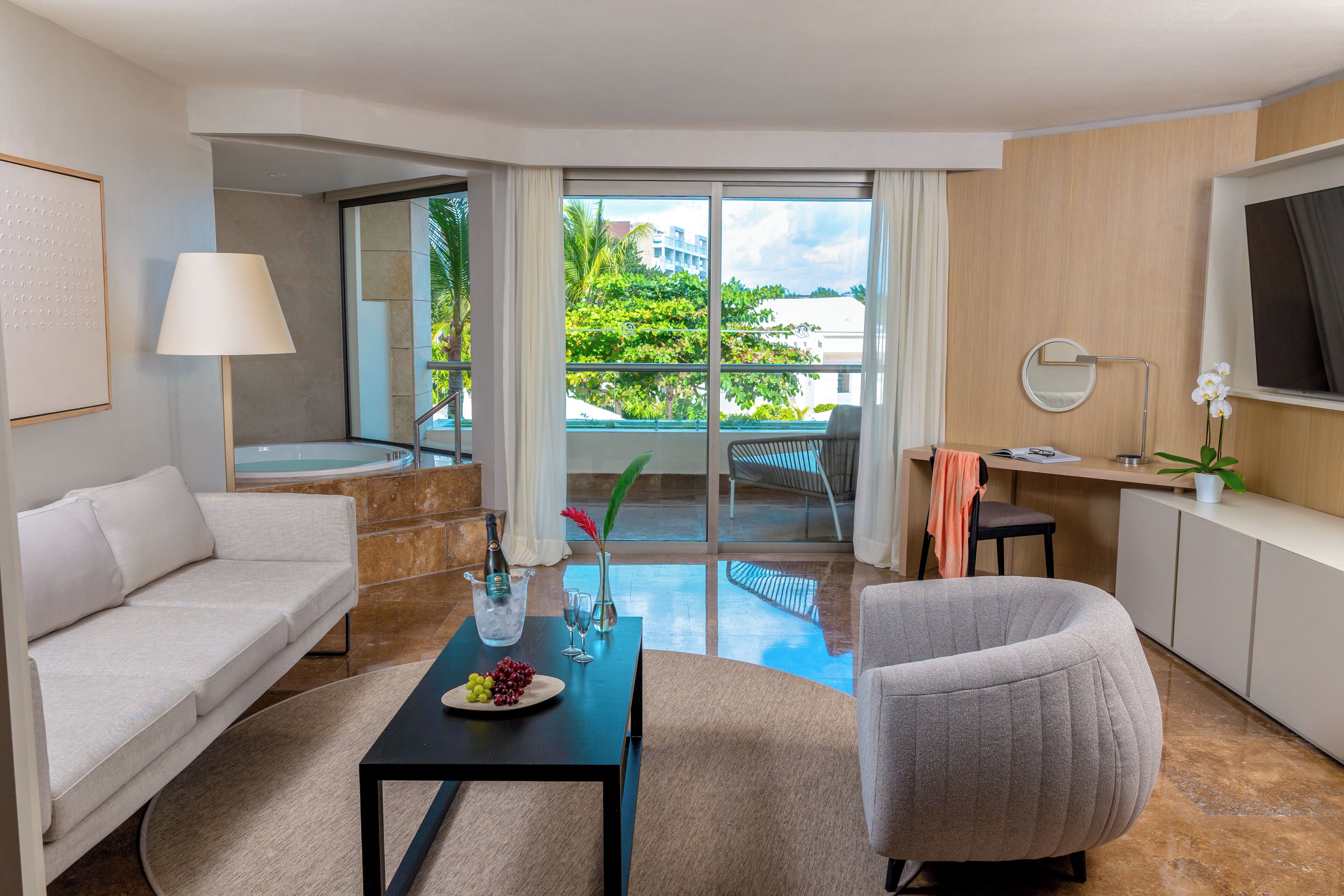 Una habitación de hotel moderna con sofá, una mesa de comedor con fruta, televisión, balcón con vistas a zonas verdes y escritorio con lámpara.