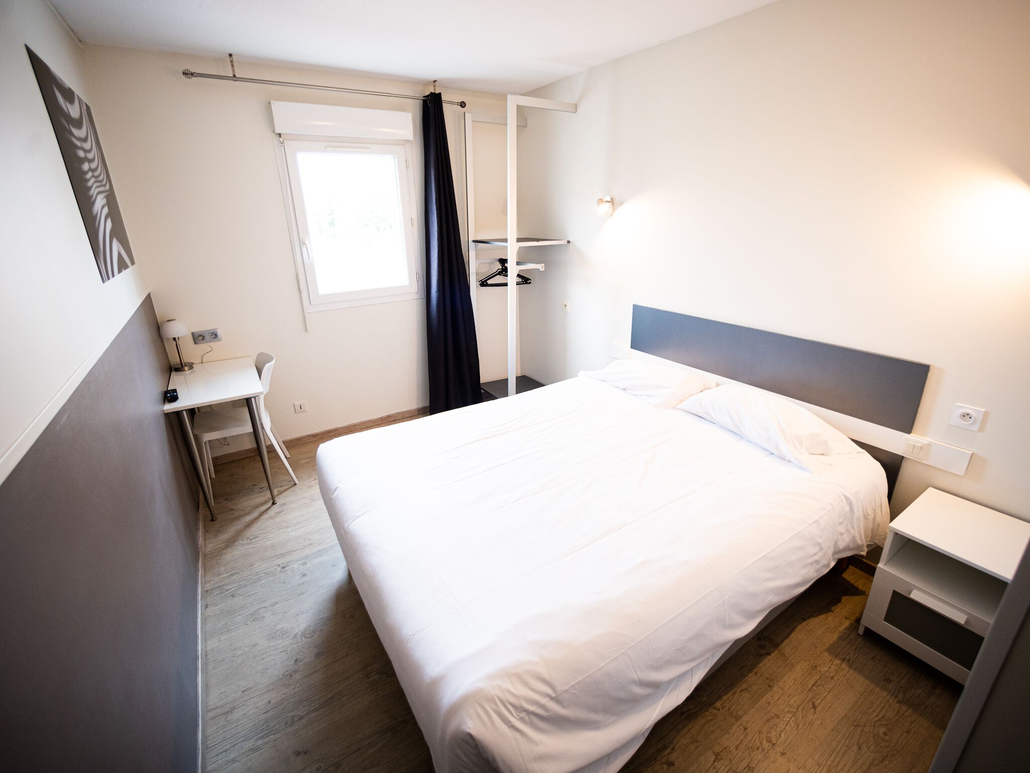 Een hotelkamer met een bed, nachtkastje, bureau, stoel en een raam met gordijnen.
