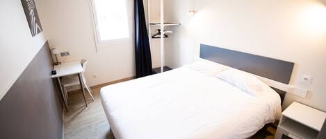 A hotel room with a bed, bedside table, desk, chair, and a window with curtains.