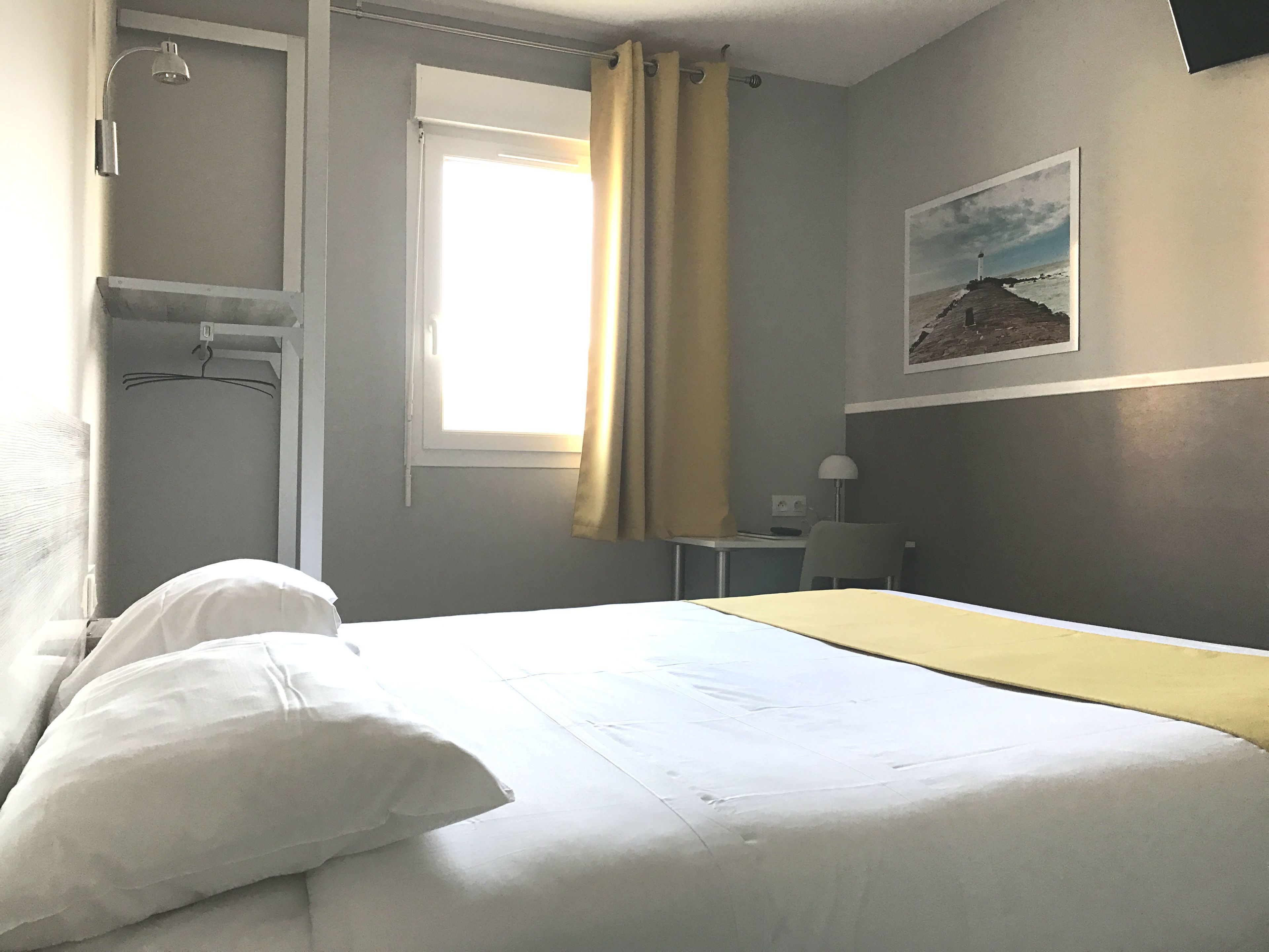 A hotel room with a bed, a desk, a chair, a window with curtains, and a wall-mounted lamp.