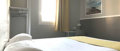 A hotel room with a bed, a desk, a chair, a window with curtains, and a wall-mounted lamp.