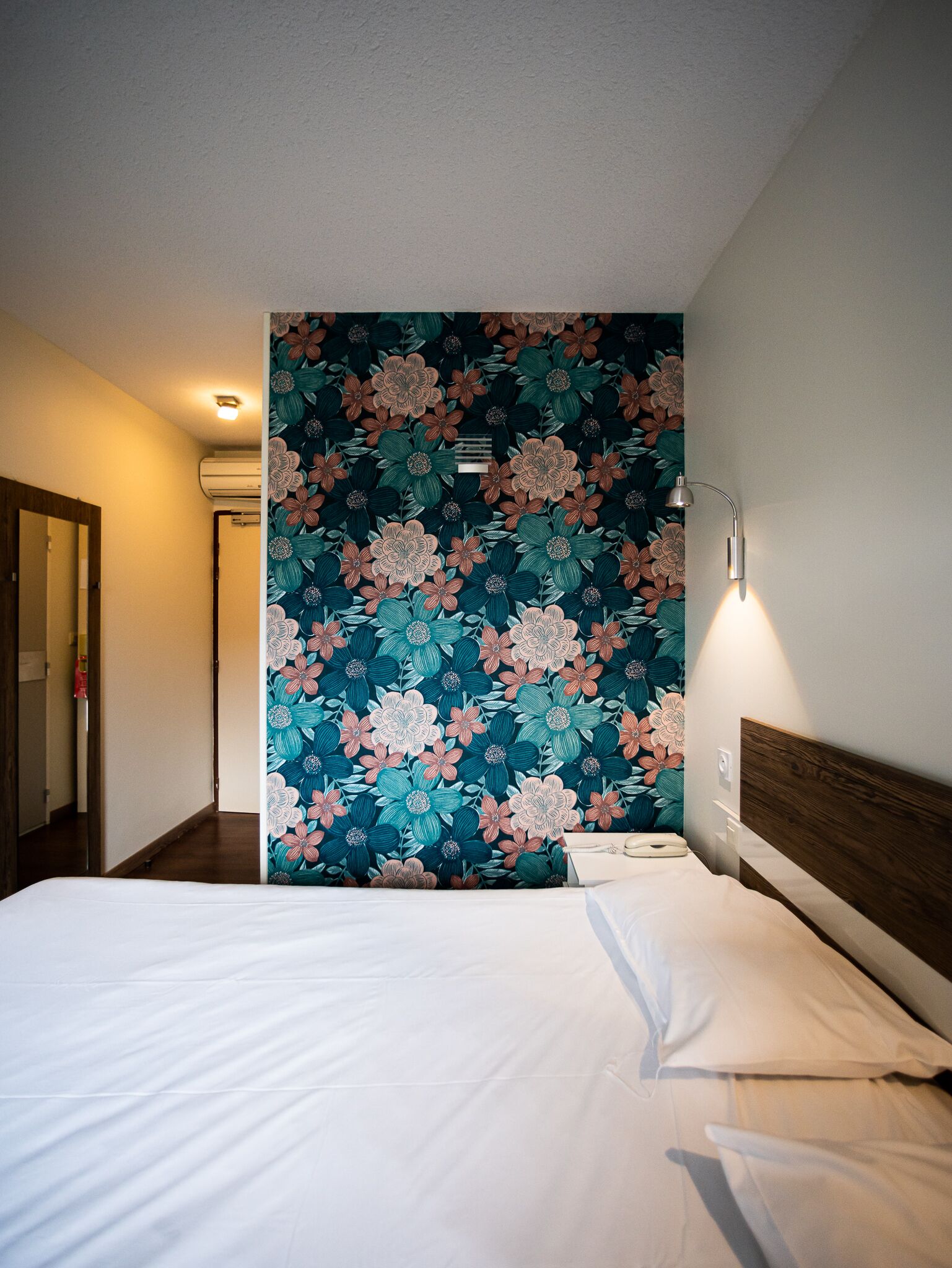 A hotel room with a large floral-patterned wall, a wooden headboard, and a bedside lamp.
