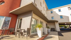 Un edificio moderno con techo plano, ventanas grandes y una zona de estar exterior cubierta con sillas y plantas en macetas.