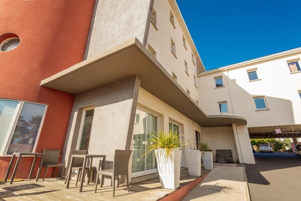 Un edificio moderno con techo plano, ventanas grandes y una zona de estar exterior cubierta con sillas y plantas en macetas.