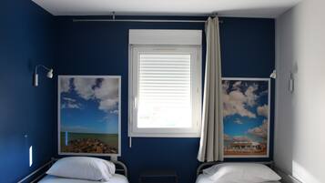 A room with two beds, a window with blinds, and two wall-mounted lamps.