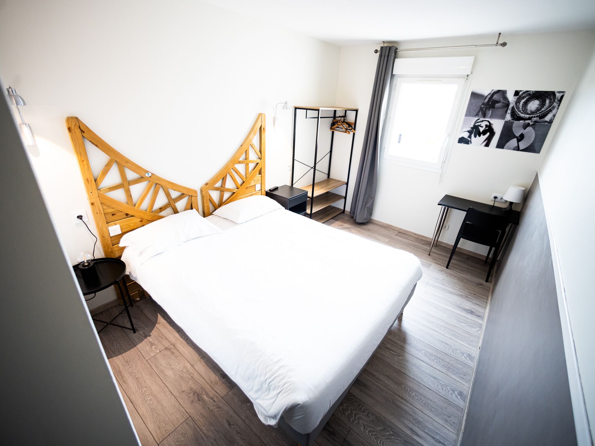 Une chambre avec une tête de lit en bois, un lit avec une literie blanche, un bureau avec une chaise, une petite table et une armoire avec une tringle pour suspendre les vêtements.