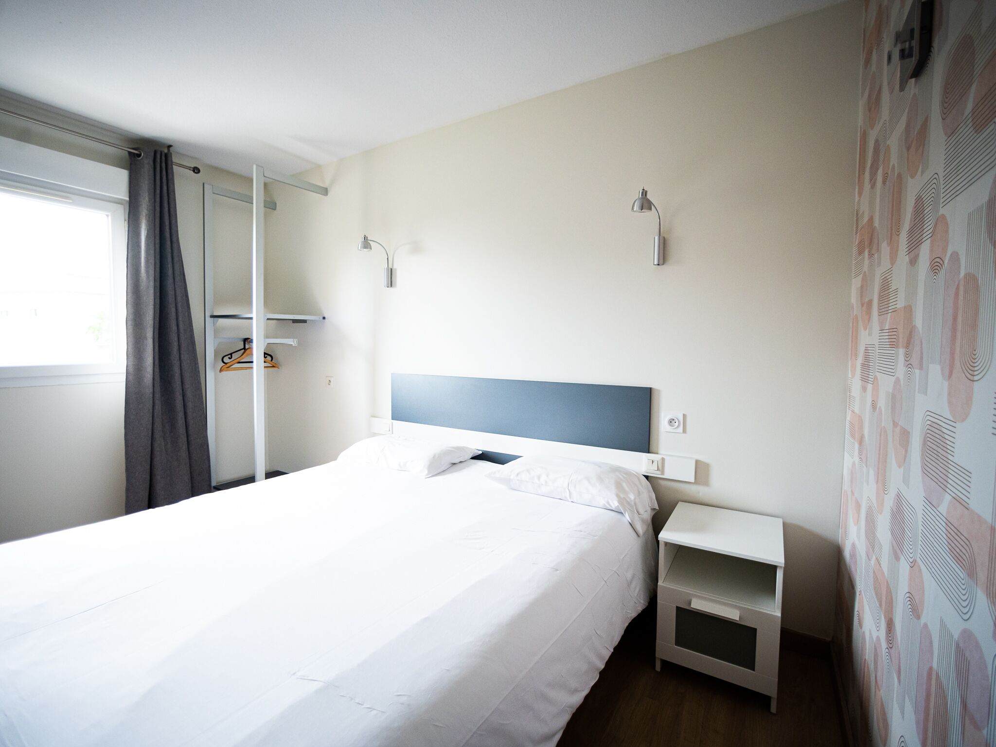 A neatly made bed in a hotel room with a white headboard, a bedside table, and a wall-mounted lamp.