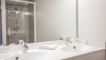 Une salle de bain avec un lavabo blanc, deux robinets, un miroir et un espace douche.
