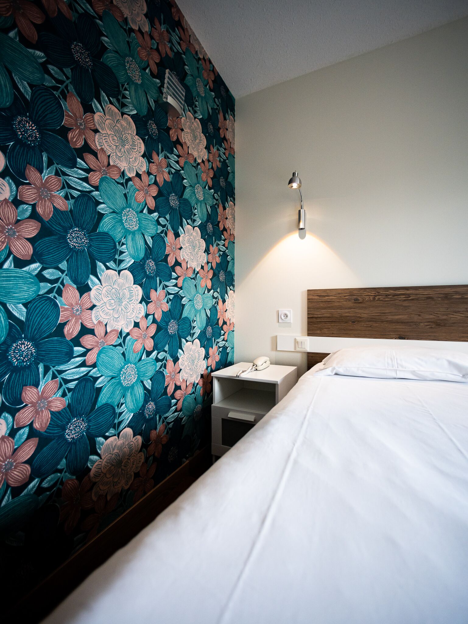 Ein Hotelzimmer mit blumigem Wandpapier, einem hölzernen Kopfteil, einem Nachttisch mit Lampe und einem weißen Bett.