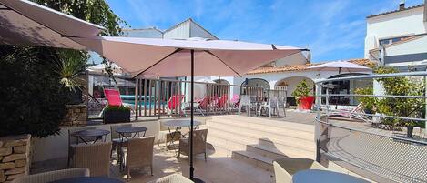 Un espace extérieur aménagé en terrasse, avec des meubles en osier, des parasols et une piscine en arrière-plan.
