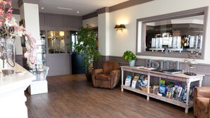 A hotel lobby with a wooden floor, a brown leather armchair, a white reception desk with brochures, a large window, and a potted plant. - Logis Hôtel de la Marée - Ile de Ré (Rivedoux-Plage)