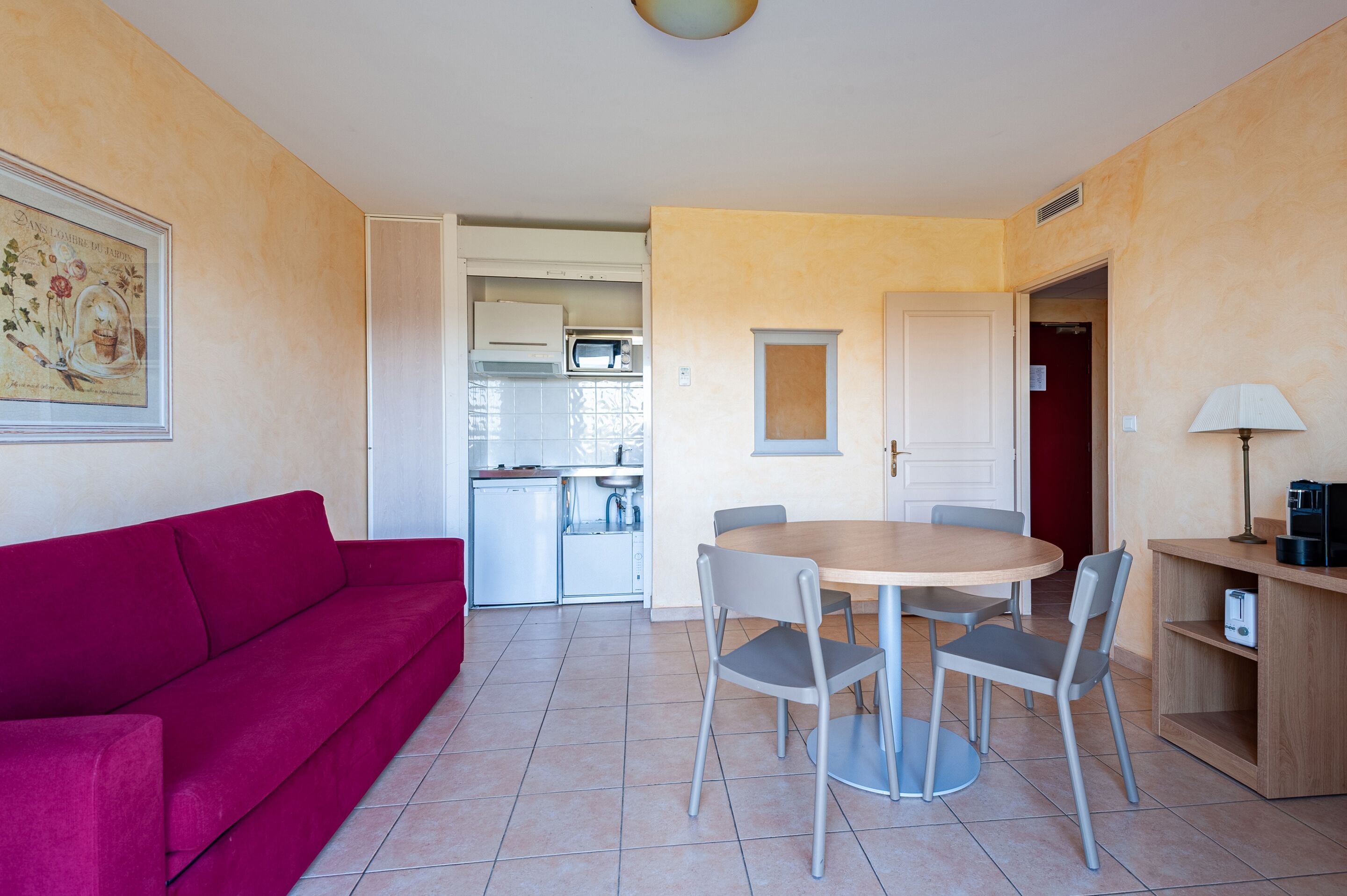 A compact living space with a red sofa, a dining table with four chairs, a kitchen area with a microwave, and a small cabinet.