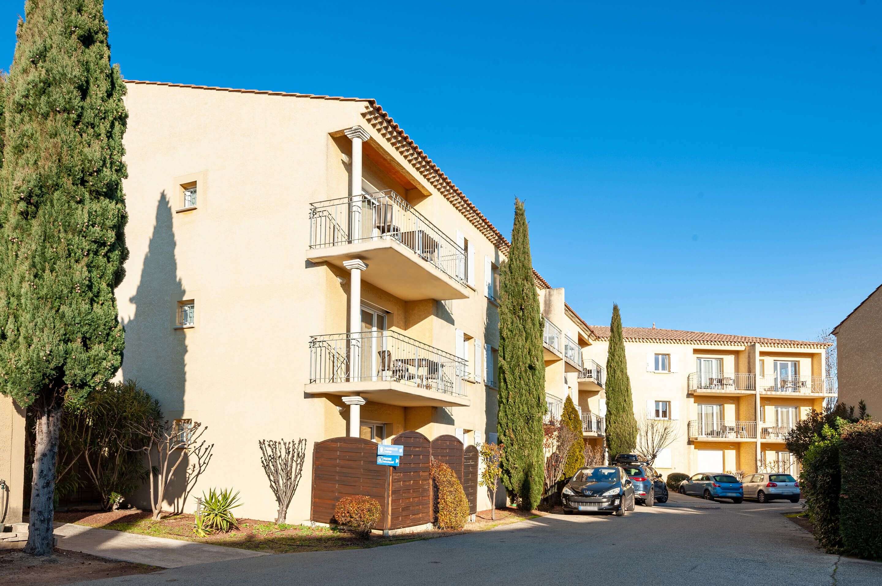 Un immeuble résidentiel à plusieurs étages, doté de balcons, d’une entrée sécurisée et de voitures garées.