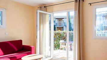 Une pièce avec un canapé rouge, une petite table en bois et une porte-fenêtre coulissante donnant sur un espace extérieur agrémenté d’un balcon et de verdure.