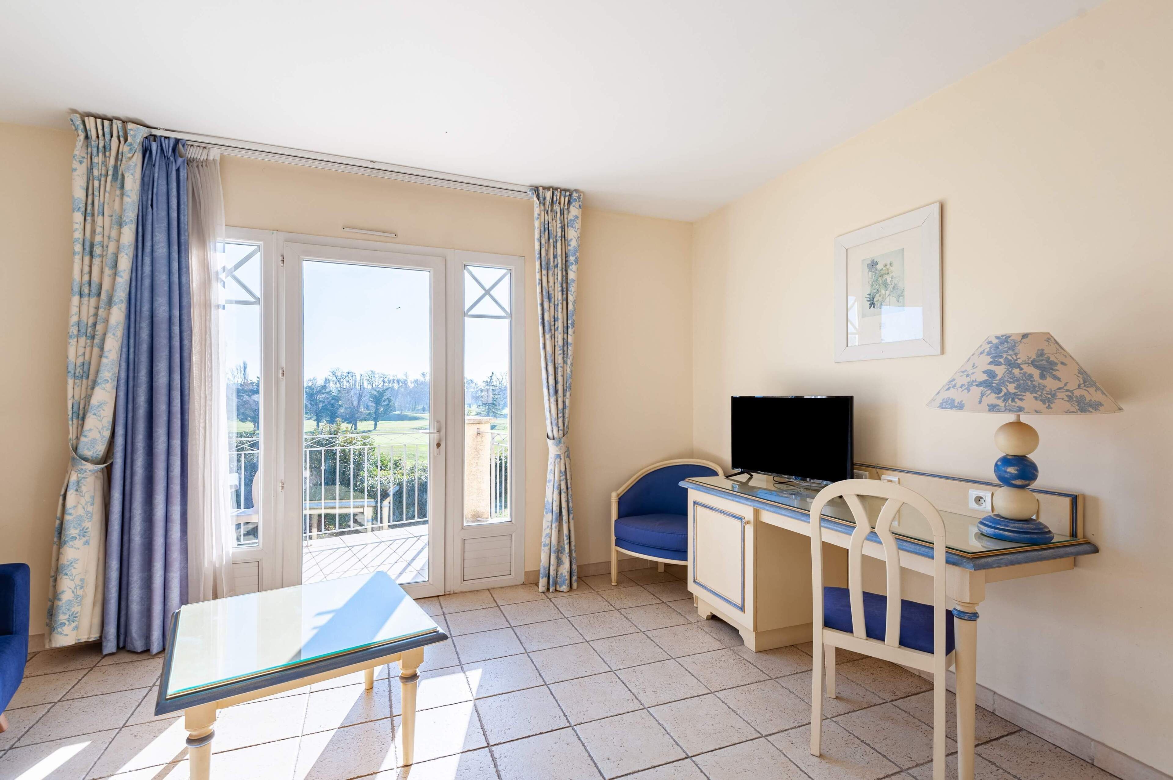 Una habitación con escritorio, silla, televisión y balcón con cortinas.