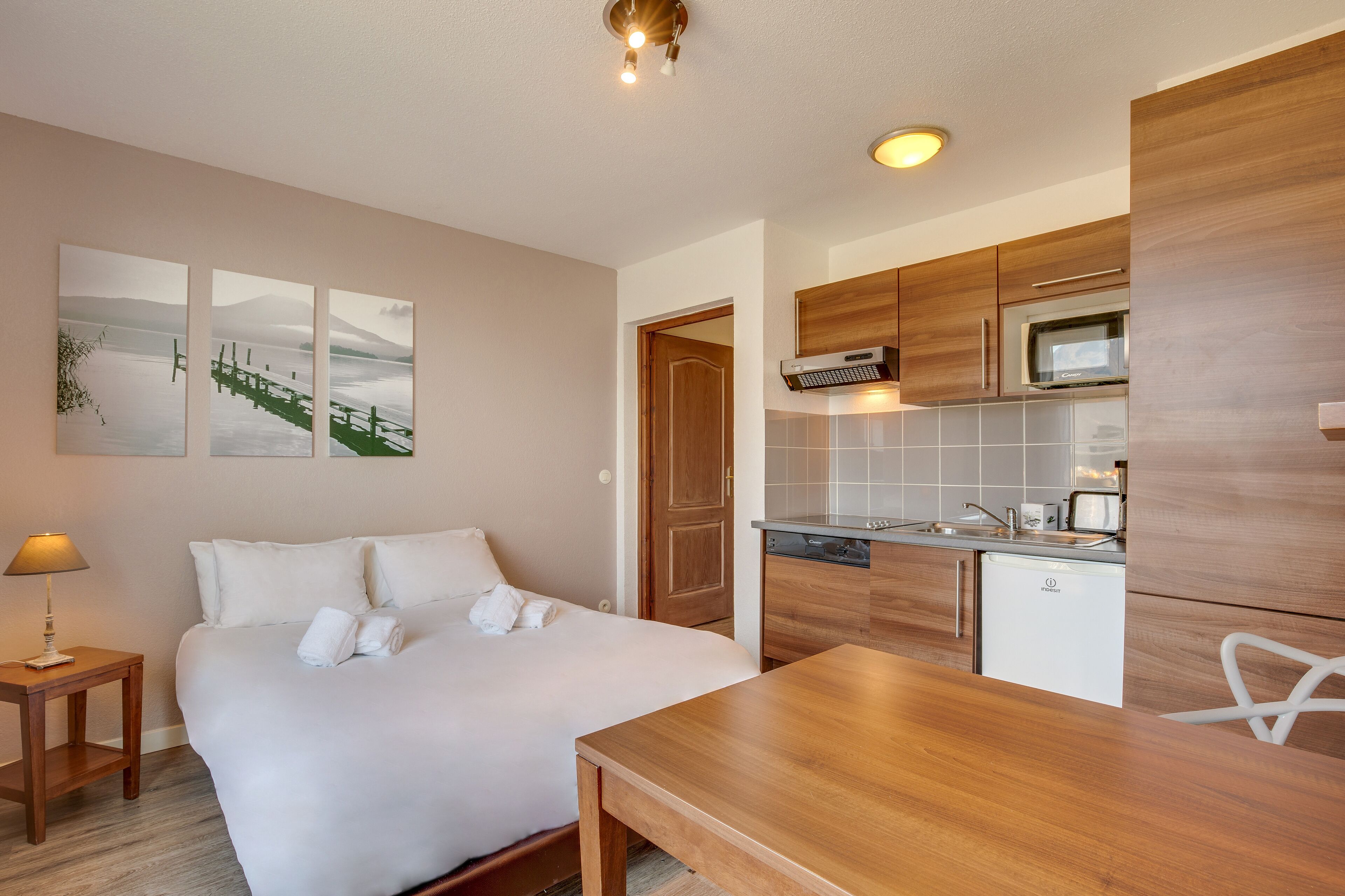 Una habitación de hotel con cama, una mesita de noche con lámpara, una mesa de comedor de madera, una zona de cocina con microondas y lavavajillas.