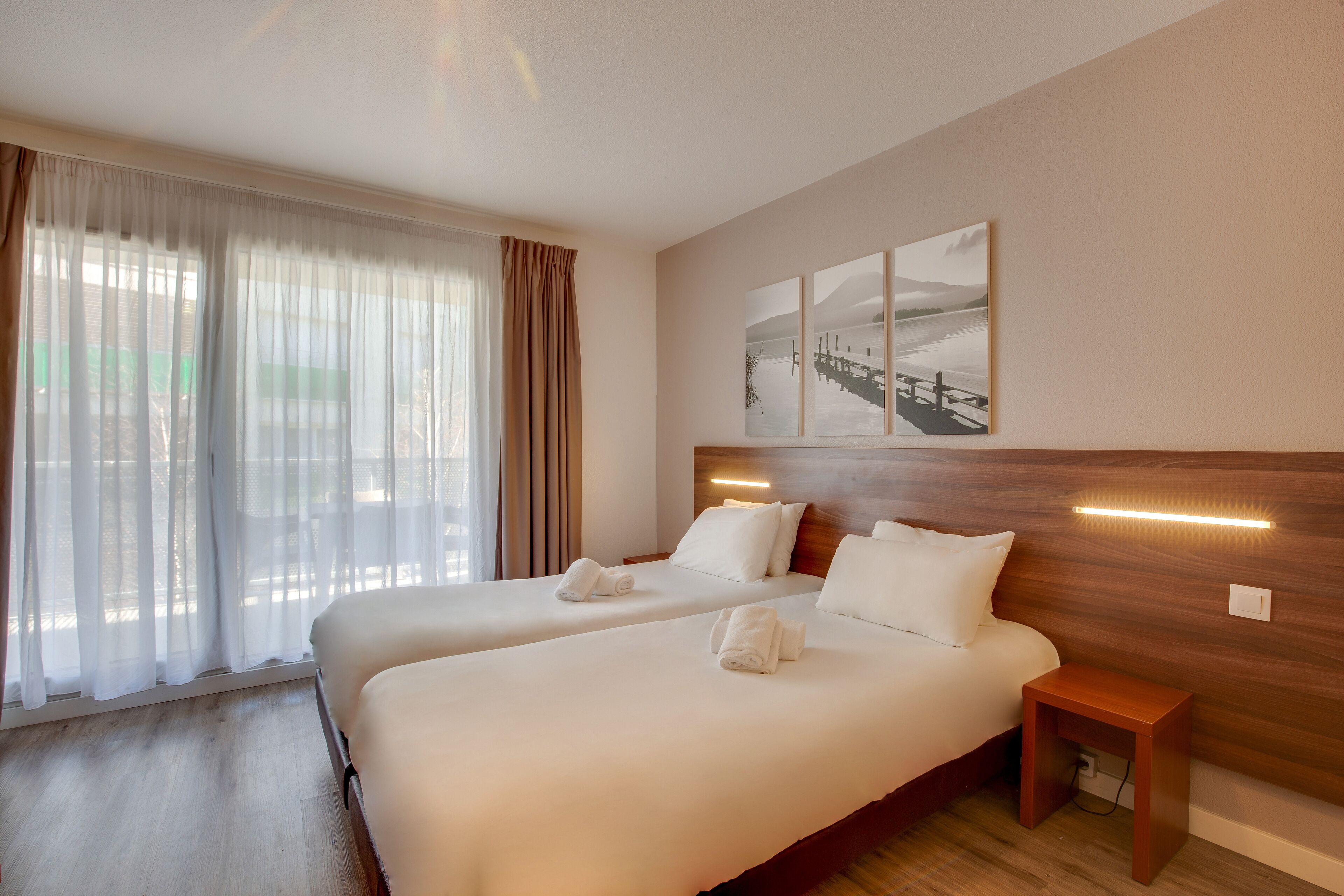 Una habitación de hotel con dos camas, cabecero de madera, una mesita de noche y una ventana con cortinas traslúcidas.