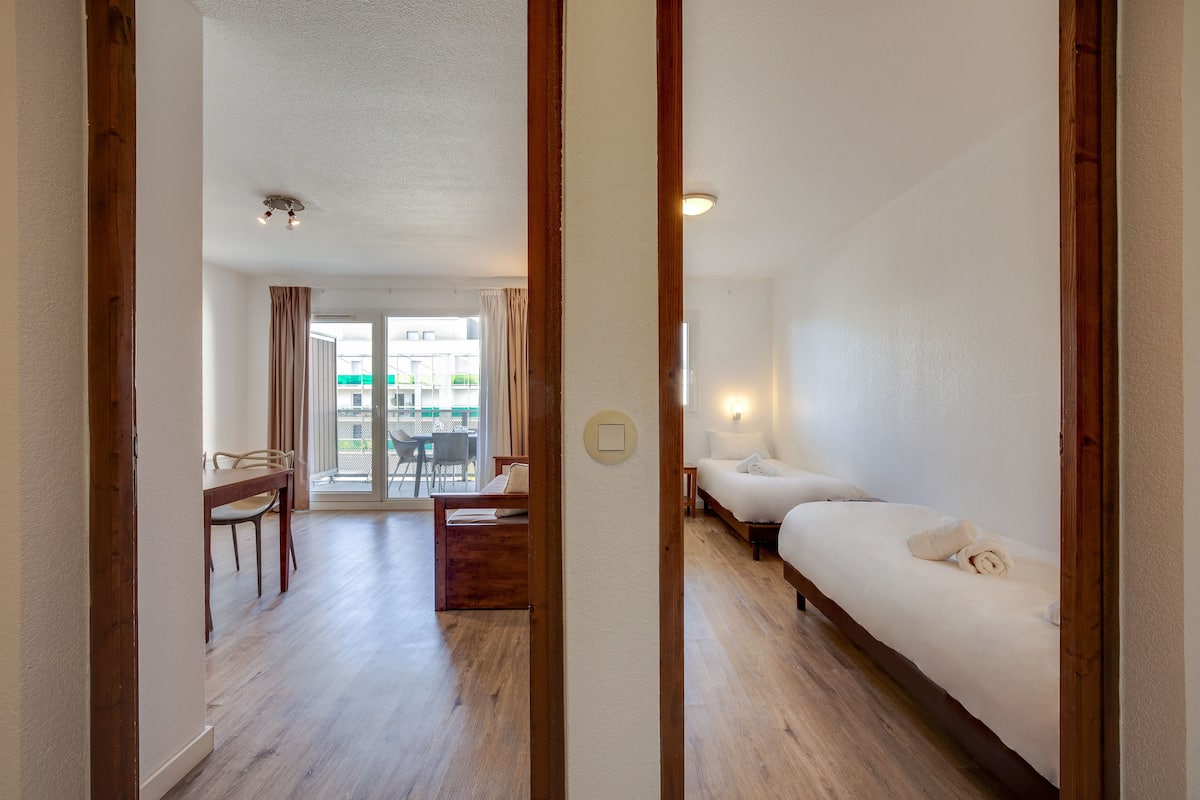 Una habitación de hotel con piso de madera, una cama con ropa blanca, un escritorio con silla y vistas al exterior a través de un vidrio.