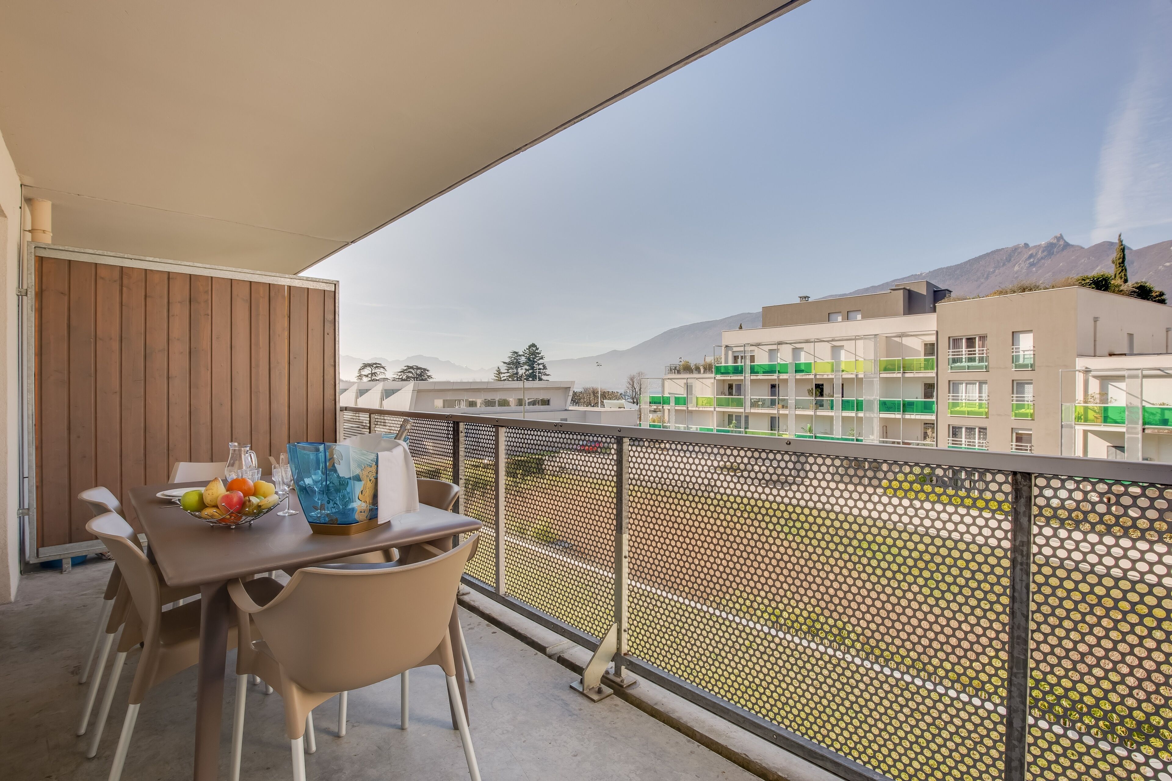 A balcony with a table set for dining, a chair, and a view of buildings and mountains in the background.