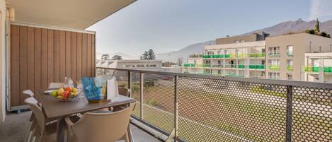A balcony with a table set for dining, a chair, and a view of buildings and mountains in the background.