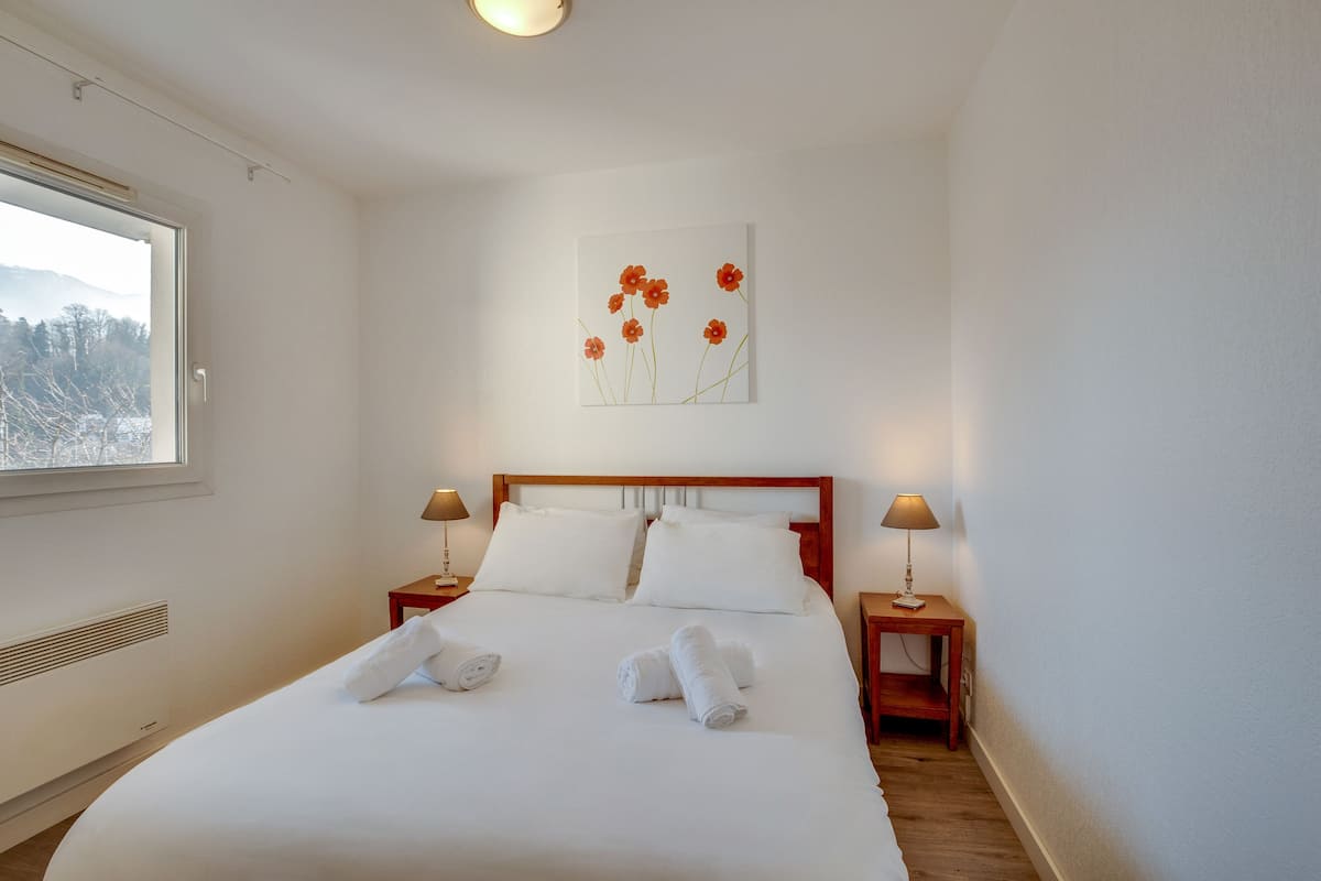 Una cama bien hecha con sábanas y toallas blancas, cabecera de madera, dos mesitas de noche con lámparas y una obra de arte de flores en la pared.