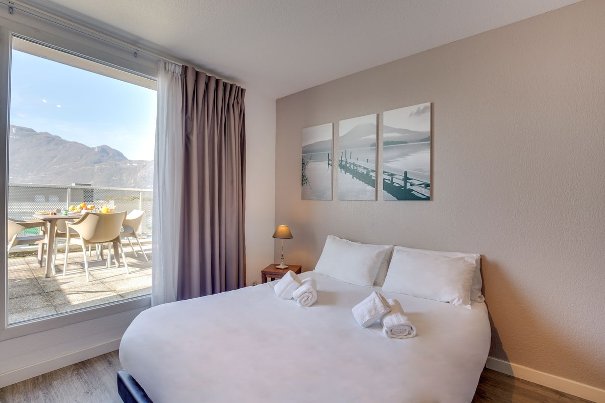 Una habitación de hotel con una cama grande, vistas a montañas a través de la ventana, una zona de comedor con sillas y una mesa, y un cuadro en la pared.