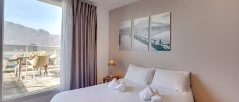 A hotel room with a large bed, a view of mountains through the window, a dining area with chairs and a table, and a painting on the wall.