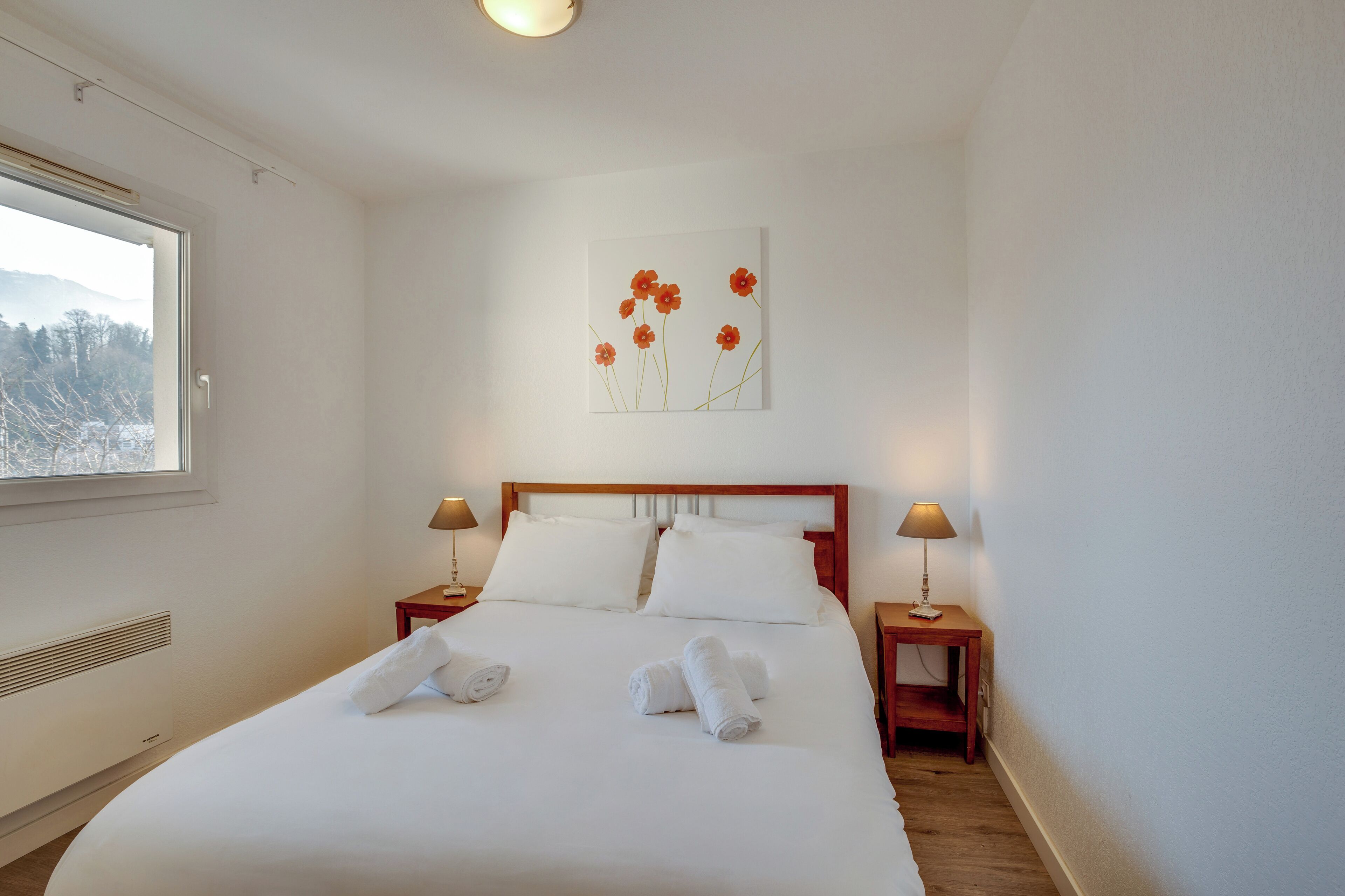 A neatly made bed with white linens and towels, a wooden headboard, two bedside tables with lamps, and a wall-mounted artwork of flowers.