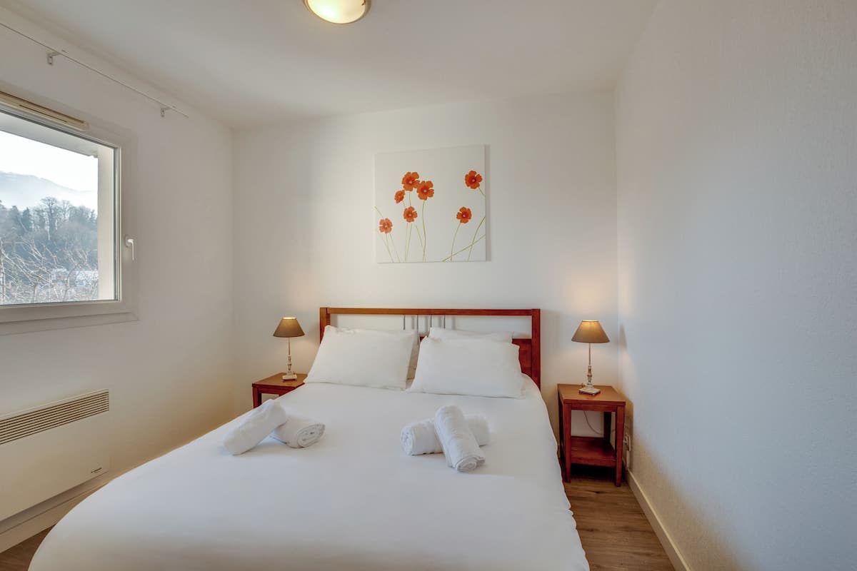 Una cama bien hecha con sábanas y toallas blancas, cabecera de madera, dos mesitas de noche con lámparas y una obra de arte de flores en la pared.